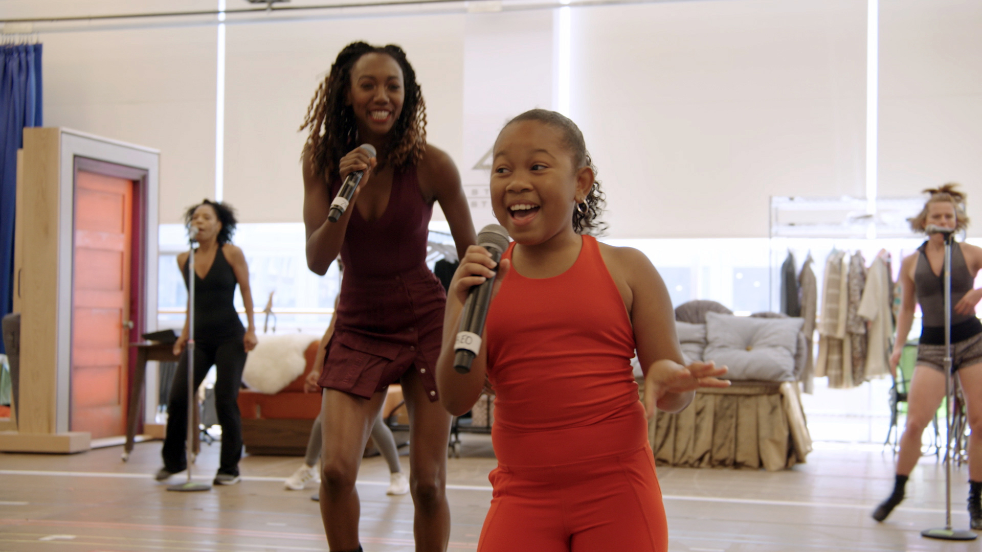 A mid-twentys woman wearing a deep purple dress stands further back, looking toward a 10-year-old in a red jumpsuit. Both are smiling and singing. Two other women are in the deep background, located in a Broadway rehearsal space.