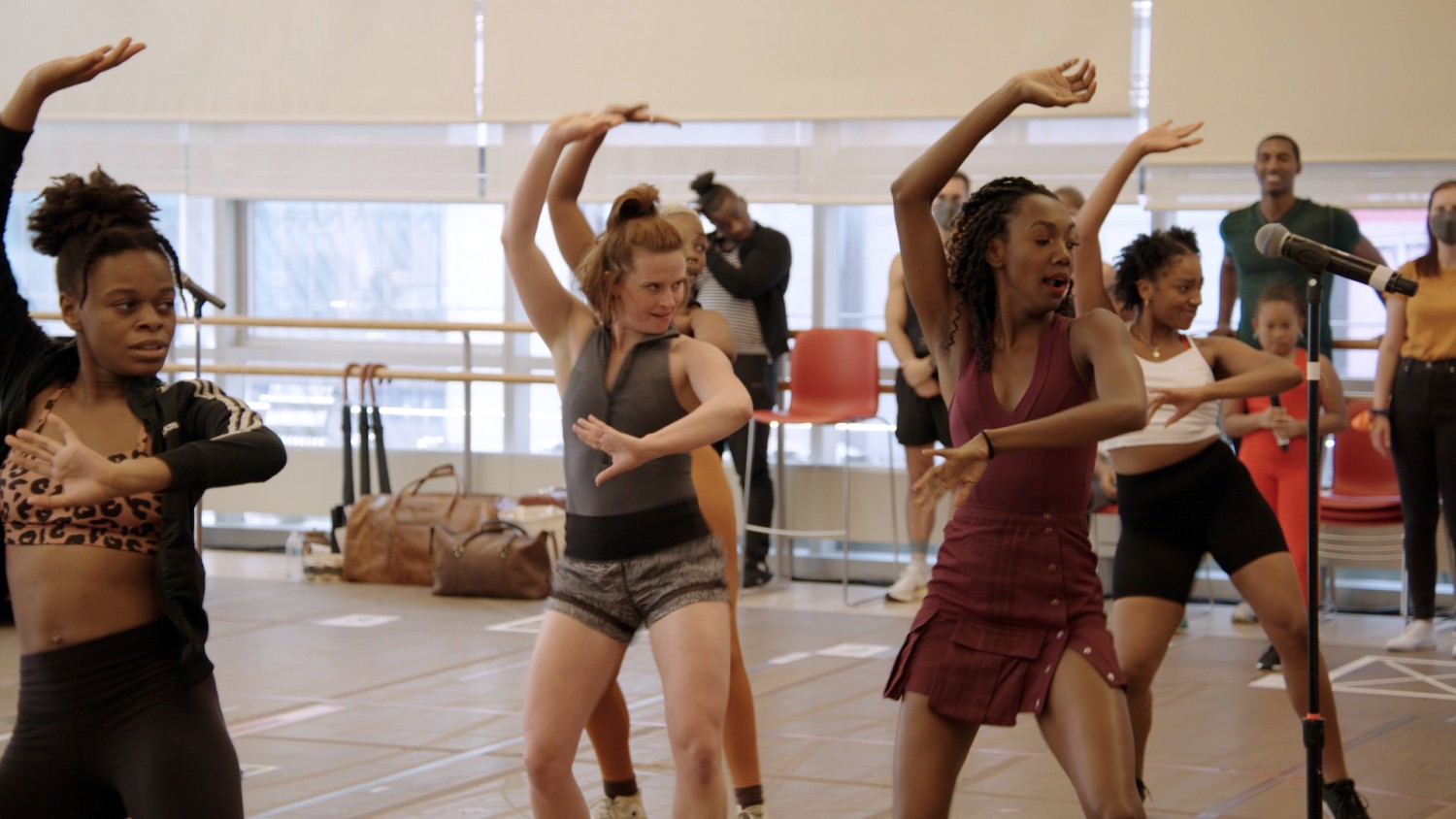A group of female Broadway performers pose in front of a microphone in a rehearsal space. Various other performers stand against windows in the background.