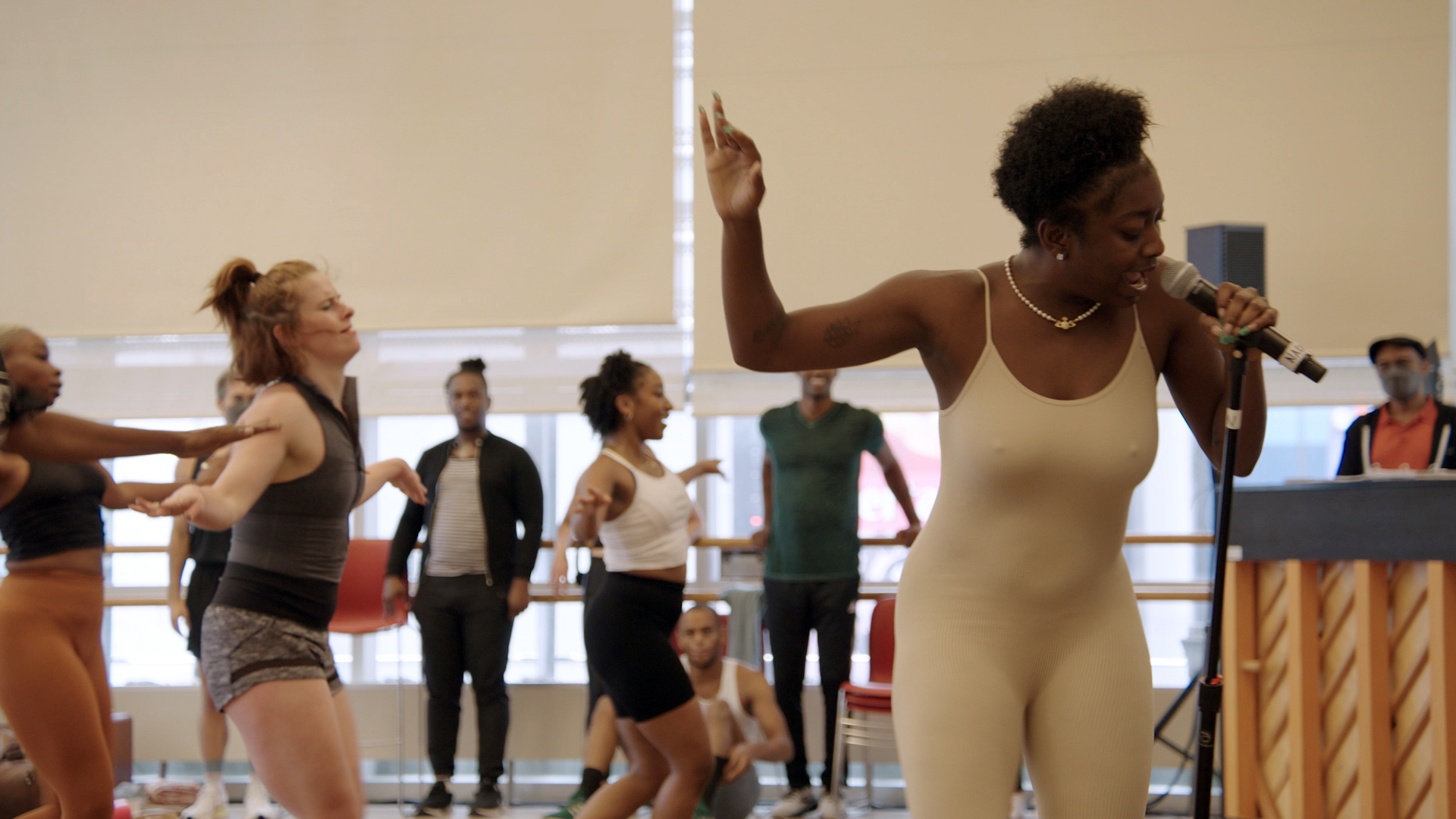 A woman in a cream bodysuit sings into a microphone on a stand, her arm raised behind her head, while dancers perform behind her. Other performers stand by the windows in the background, watching next to a piano.