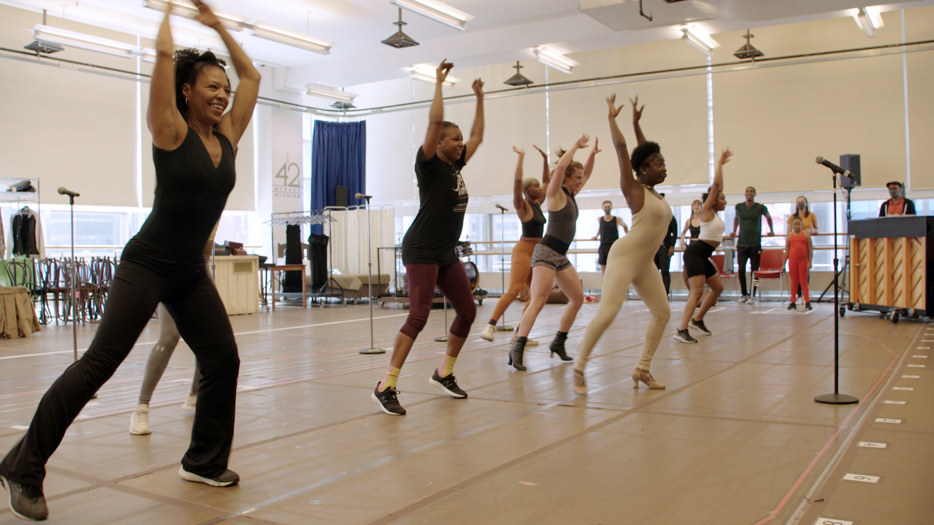 A group of female Broadway performers pose in front of a microphone in a rehearsal space. Various other performers stand against windows in the background.