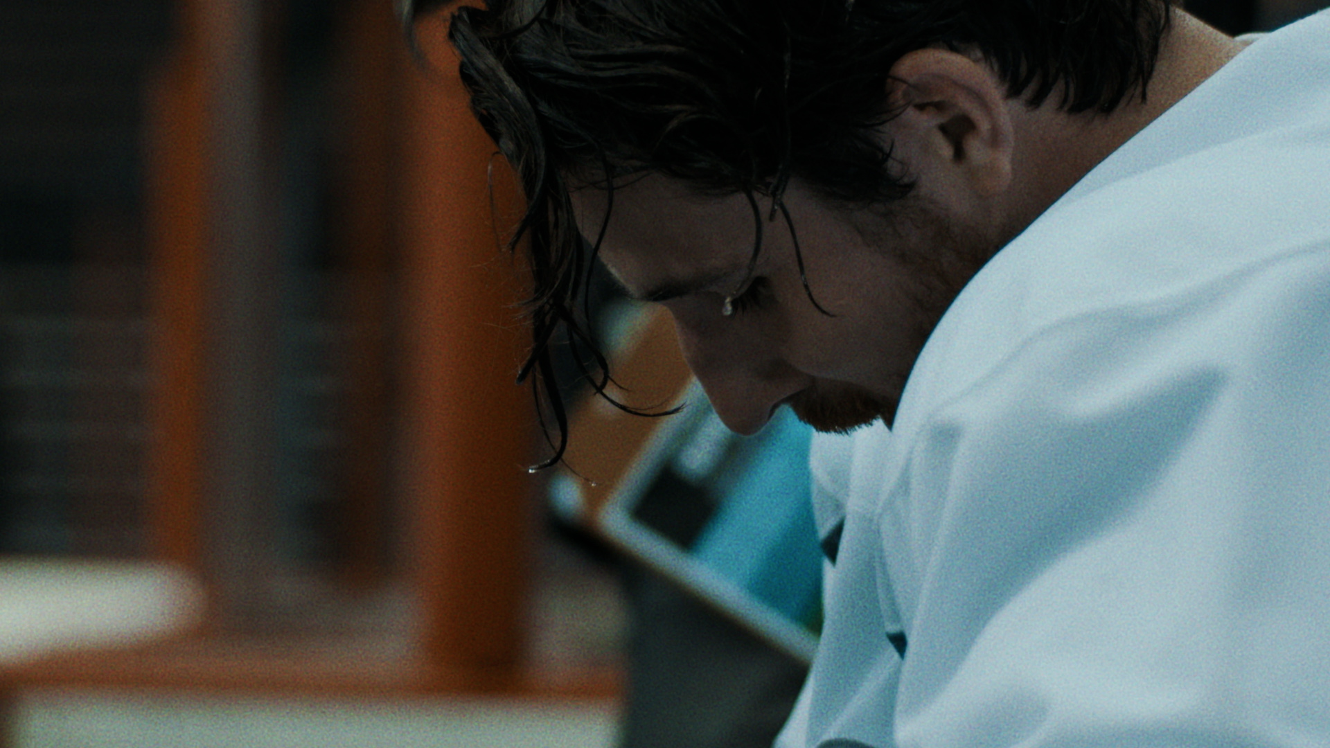 A hockey player in a white jersey sits on the bench. Beads of sweat drip from long strands of hair dangling in front of his face.