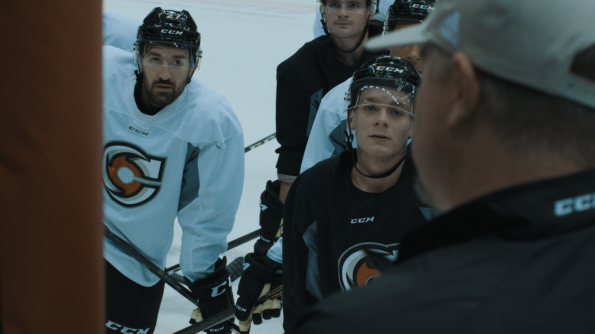 Hockey players wearing white and black jerseys take a knee on the ice, looking up at a coach who is in the foreground. His face is obscured, but he wears a black jacket and cream-colored hat. The logo for the Cincinnati Cyclones is on the jerseys.