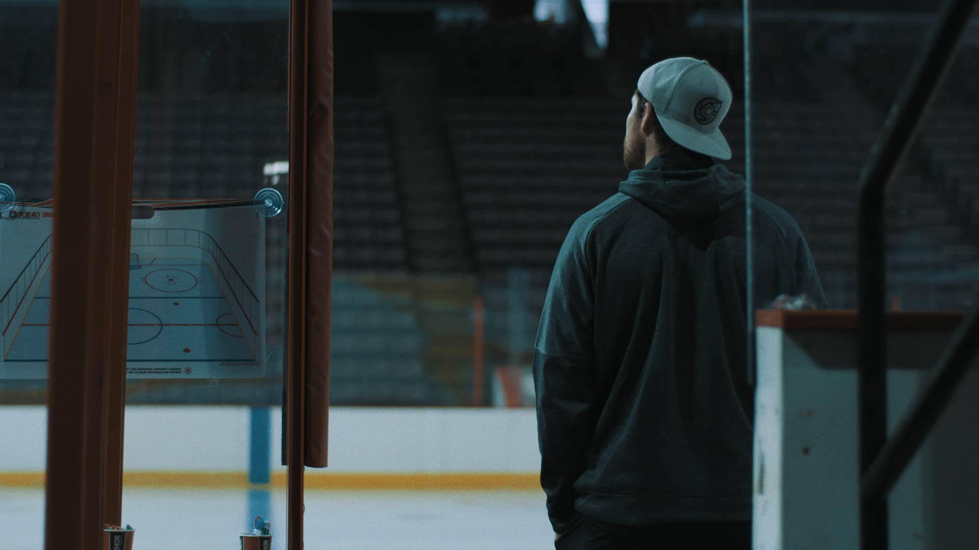 A man stands at the edge of a hockey rink, looking to the distance. He wears a grey hoodie and white backwards cap and is facing away from the camera. The arena is empty, and a coach’s dry erase board is hung on the glass.
