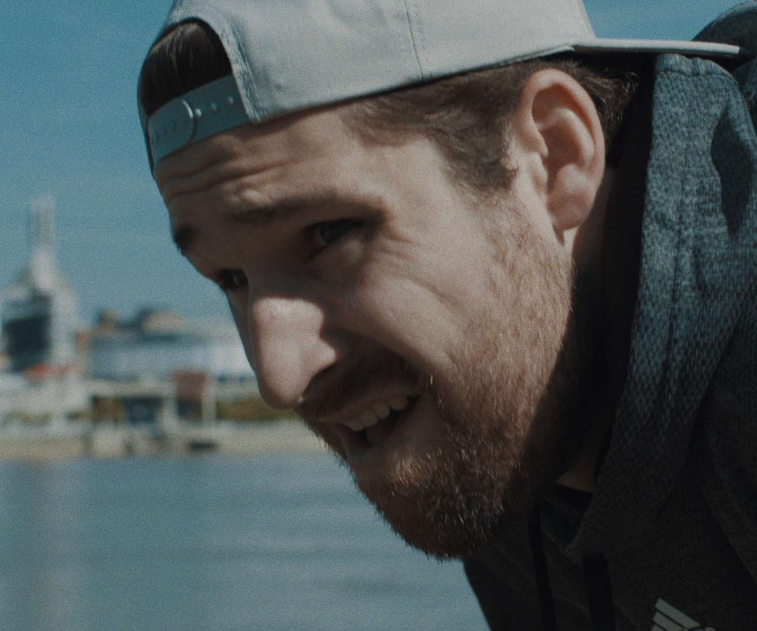 A man in a grey Adidas hoodie and white backwards cap stands hunched over in front of a riverbank. He is catching his breath.