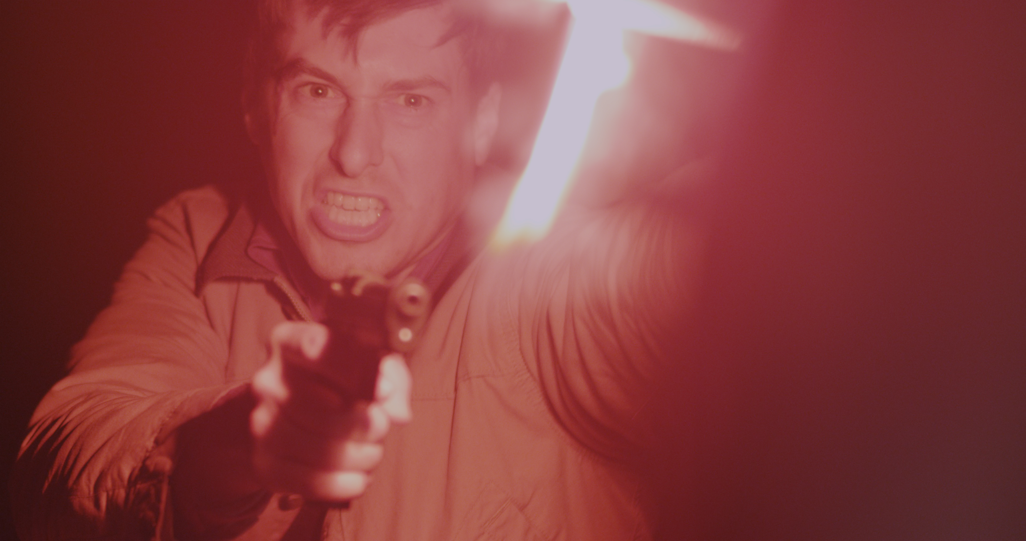 An angry man, wearing a tan jacket, points a pistol to the side of the camera. He holds a lit road flare.