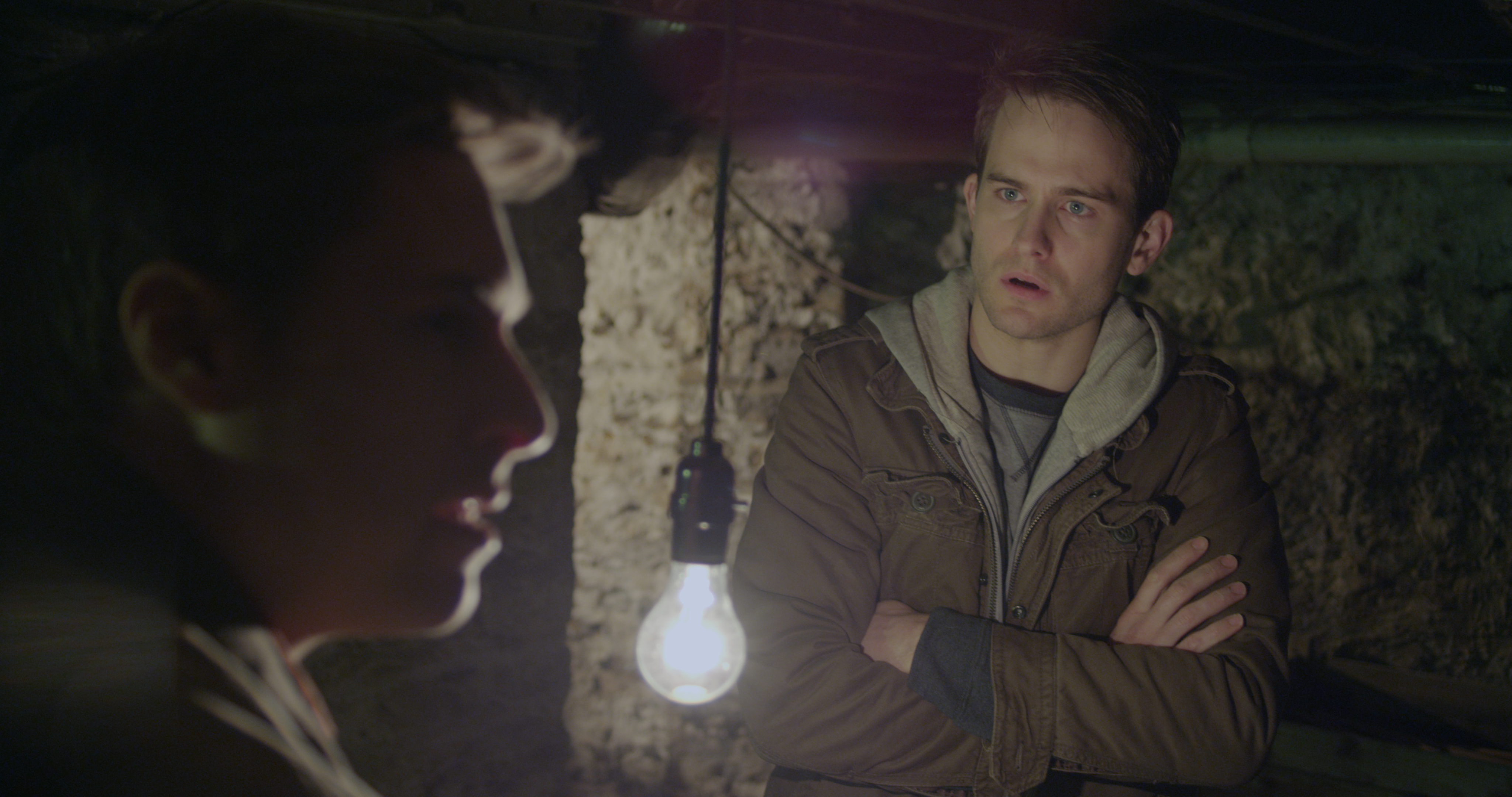 A man in a brown jacket and grey t-shirt looks stunned at another man in the foreground. The second man is out of focus and looking off to the side. A single lightbulb hangs between them in an unfinished basement.
