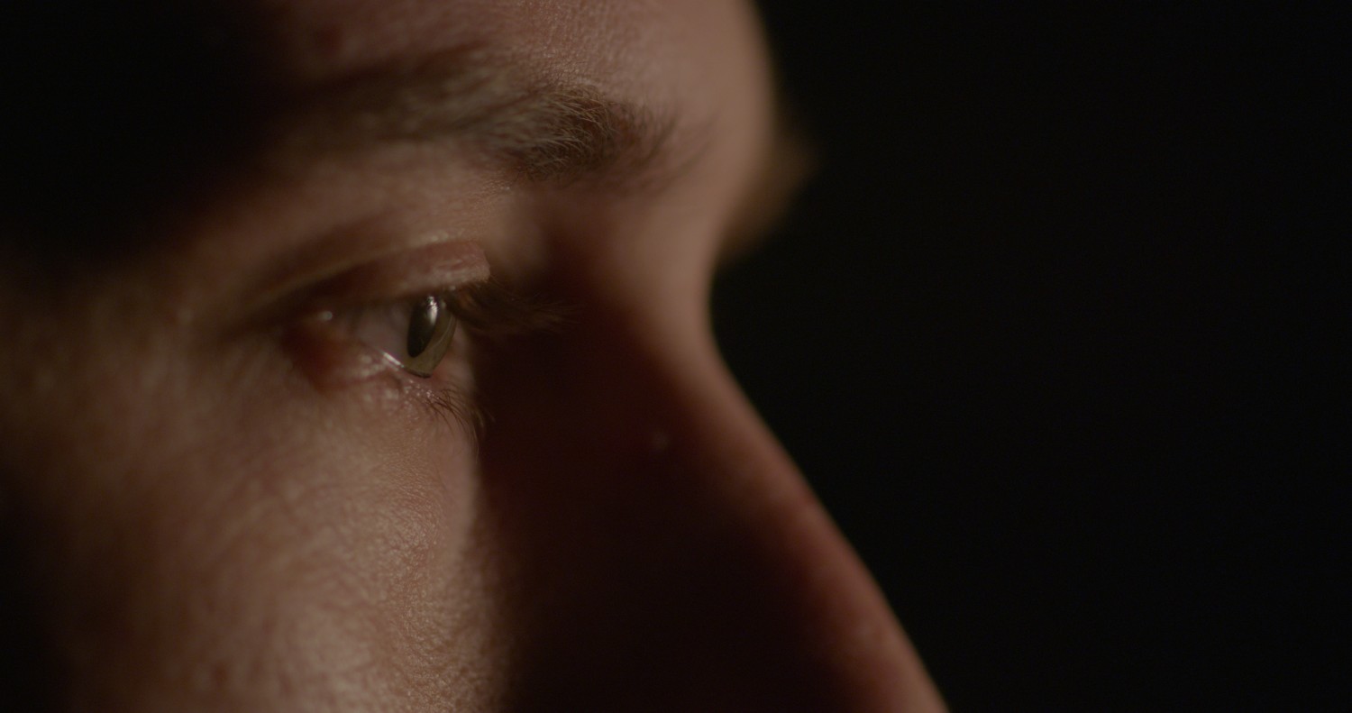 Macro lens shot of a man’s eyeball. He looks from screen left to right, and there is a bright light hitting his face directly from the front. The background is completely black.