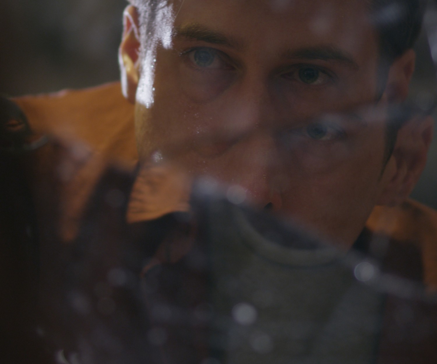 Man in a deep brown shirt looks at himself in a broken mirror. The shattered glass reflects multiple faces back at him.
