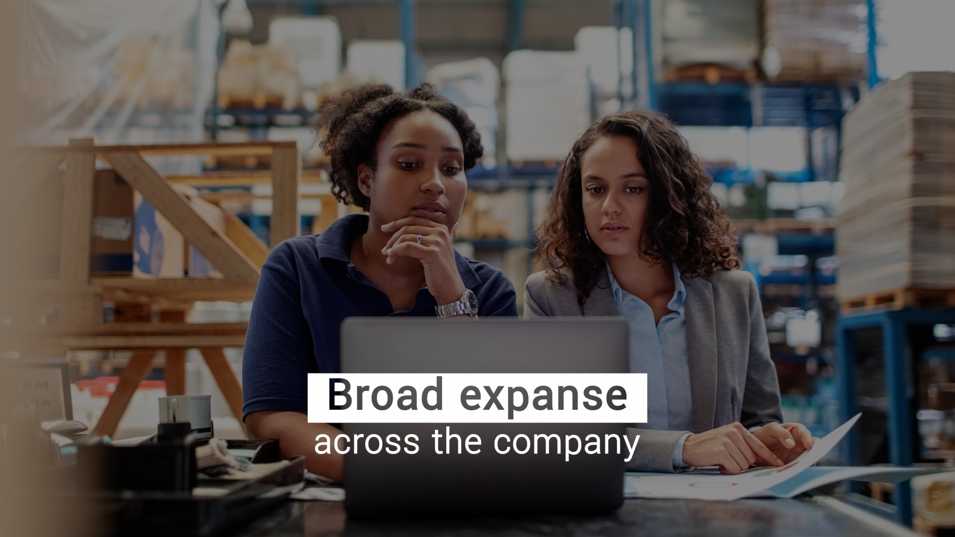 Two women stand at a desk in a warehouse, looking at a laptop. The words “Broad expanse across the company” hover in front of the laptop in white text.
