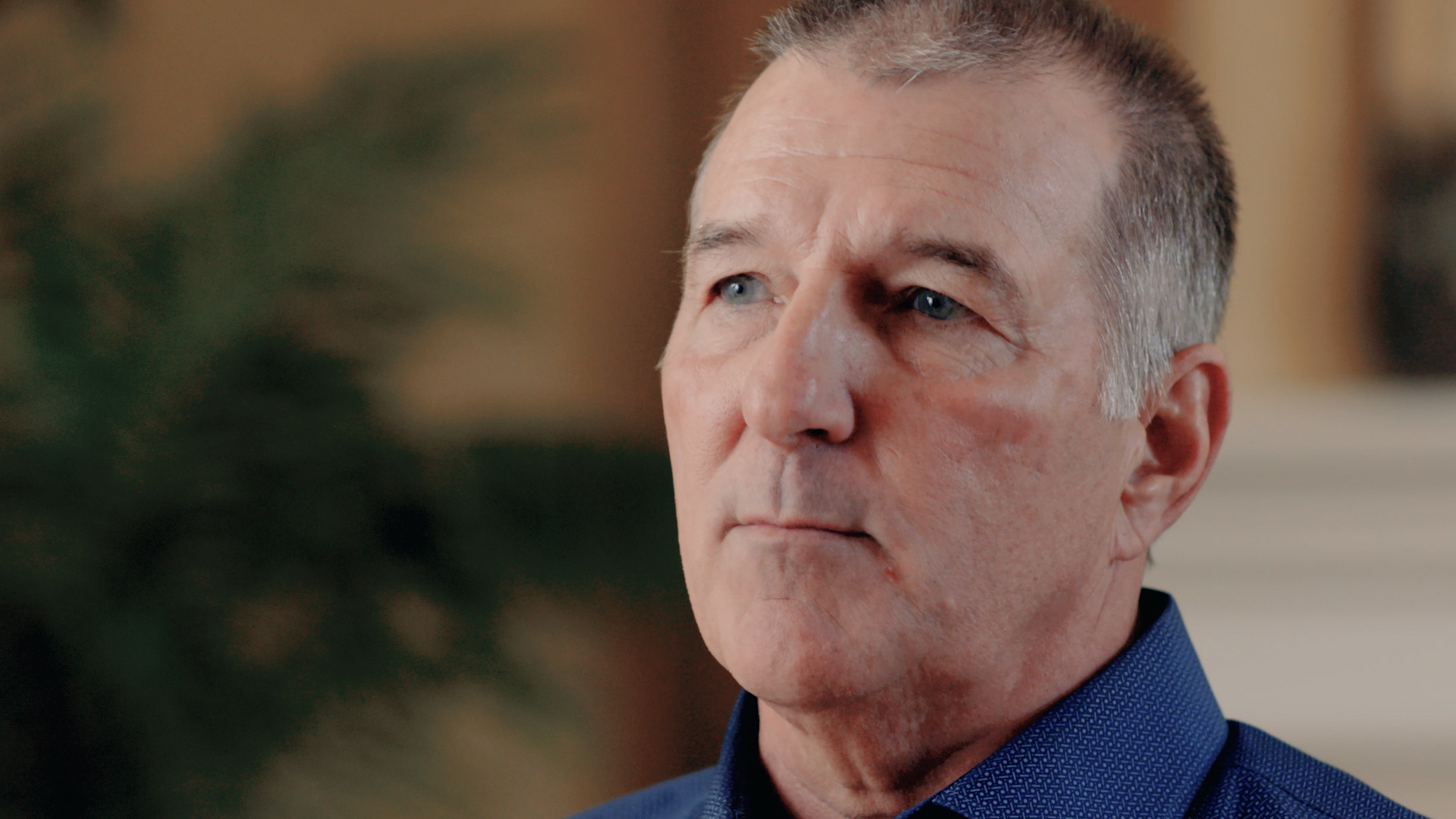 Mike Racer is interviewed in a living room, seated in front of a white fireplace and one plants. He wears a blue button up shirt.