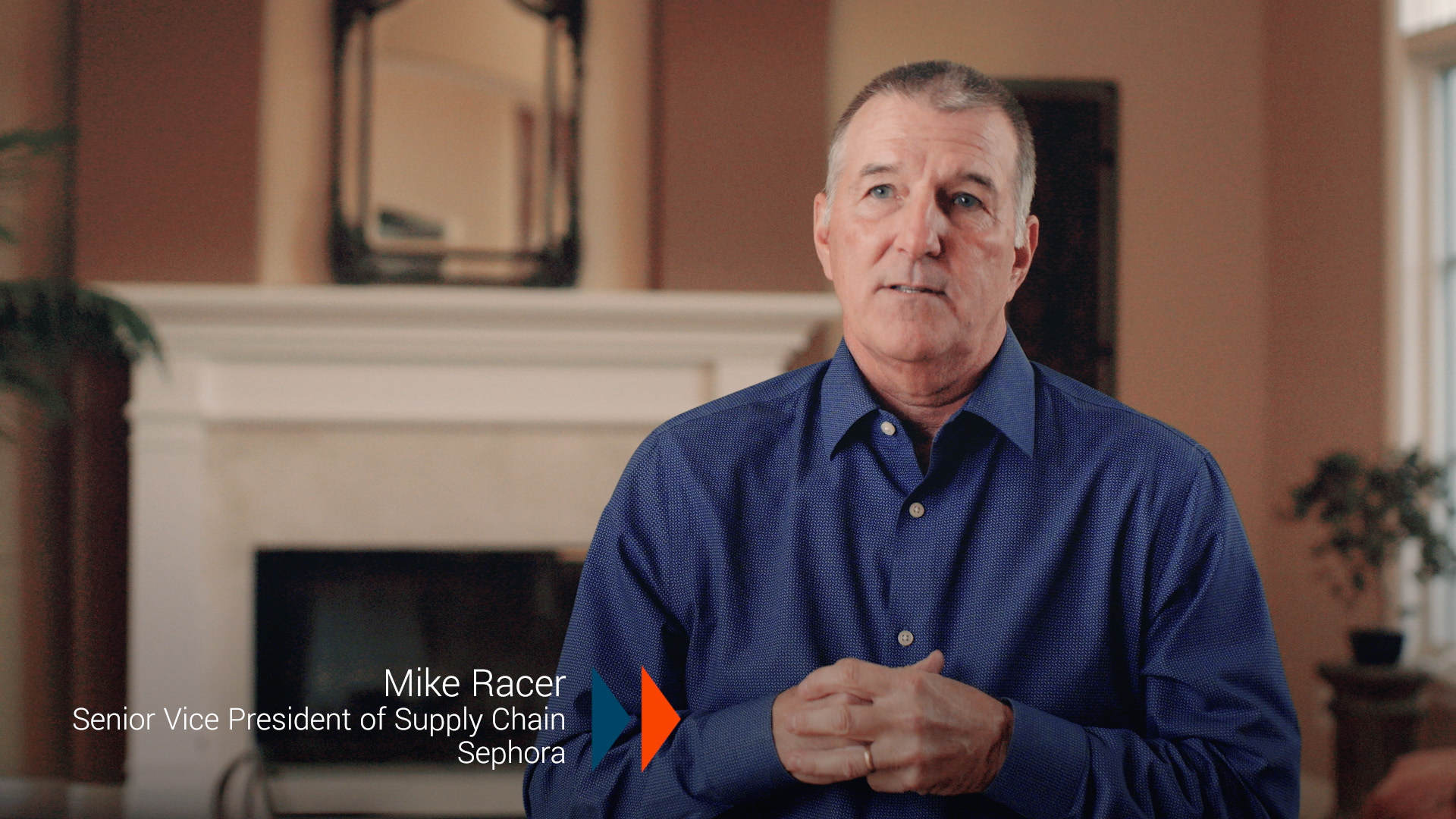 Mike Racer, Senior Vice President of Supply Chain for Sephora is interviewed in a living room, seated in front of a white fireplace, mirror and two plants. He wears a blue button up shirt and has his hands clasped in front of his chest. His name and title appear in a lower third graphic.