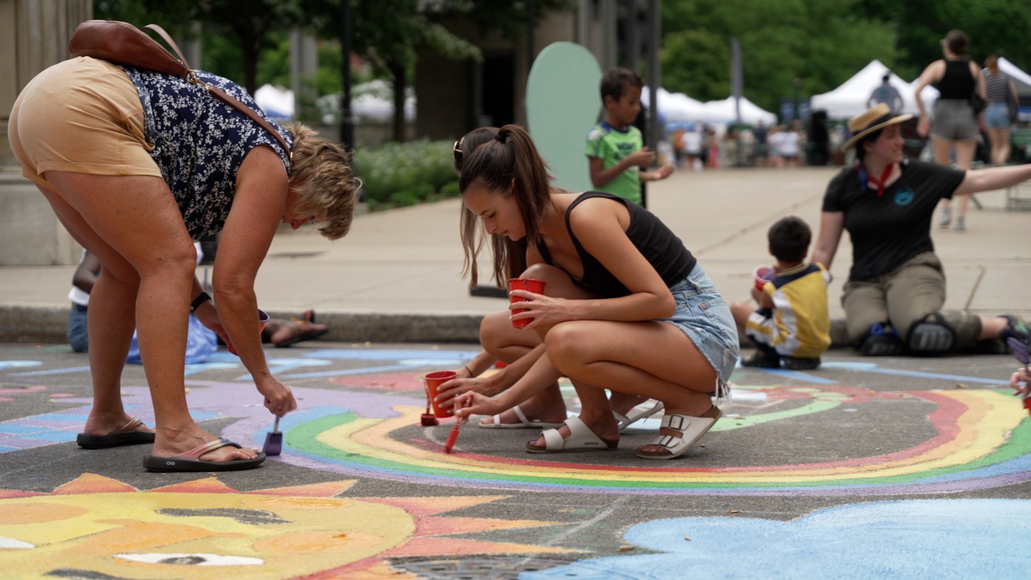Two women bend down and paint a rainbow on a street, in the middle of a mural. The two are surrounded by others working on the same project.