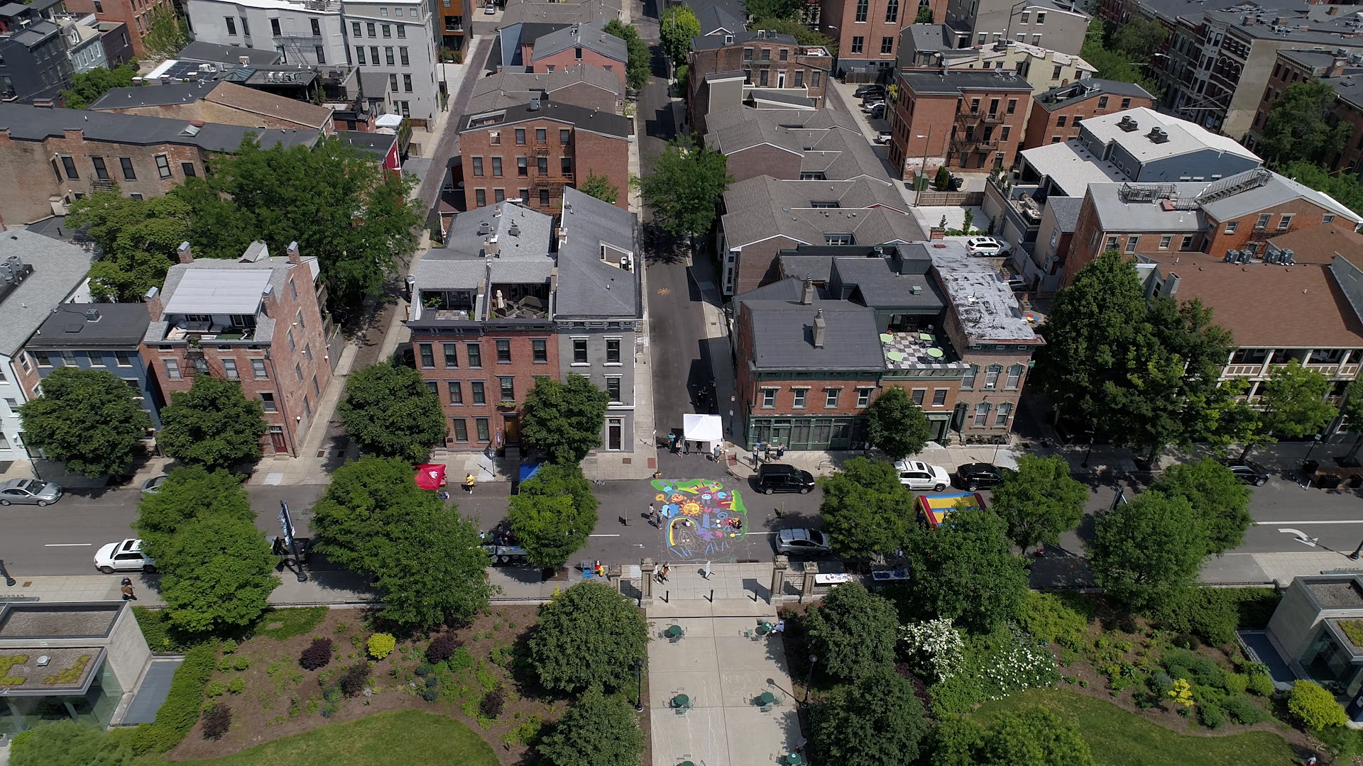 An aerial view of OTR in Cincinnati, featuring a fresh mural painted at the intersection of two streets. Artists complete the finishing touches on a sunny day.