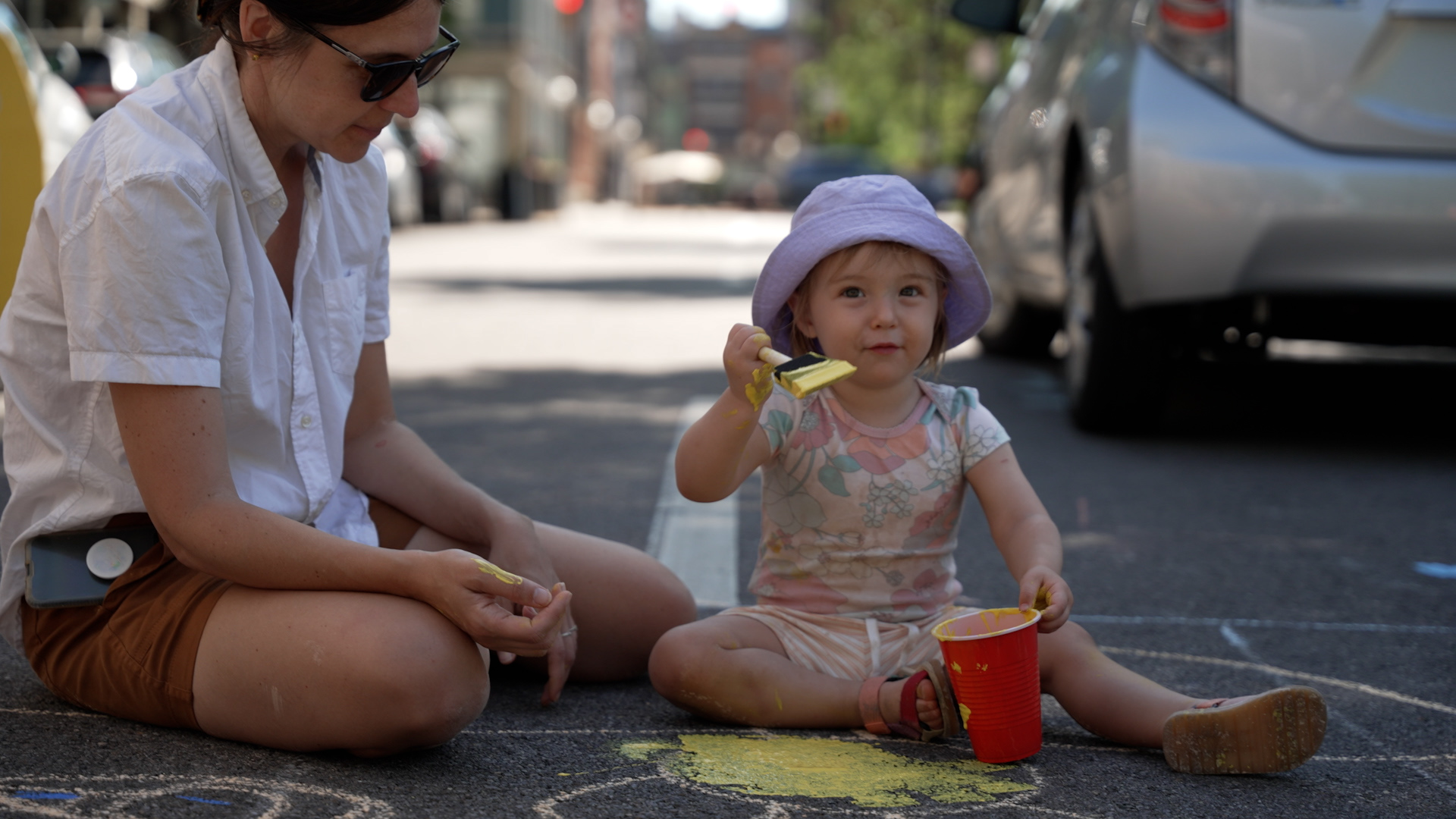 A mother and daughter sit in the road, working on a street painting. The young girl wears a purple hat and floral jumper and sandals and holds a red solo cup filled with yellow paint in one hand and a paintbrush in the other. Her mother wears a white shirt and brown shorts.