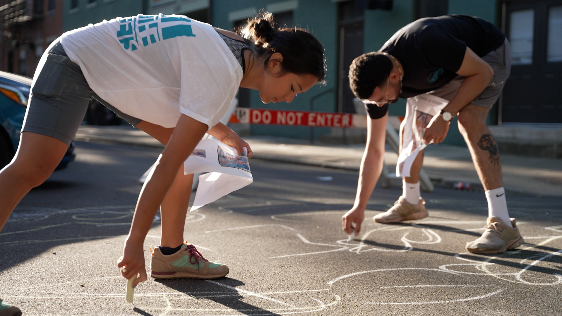 A woman in a white t-shirt, grey shorts and gym shoes and man in black t-shirt, grey shorts and gym shoes bend over and use chalk to draw figures on the pavement. They each hold a piece of paper with the plans for the art included.