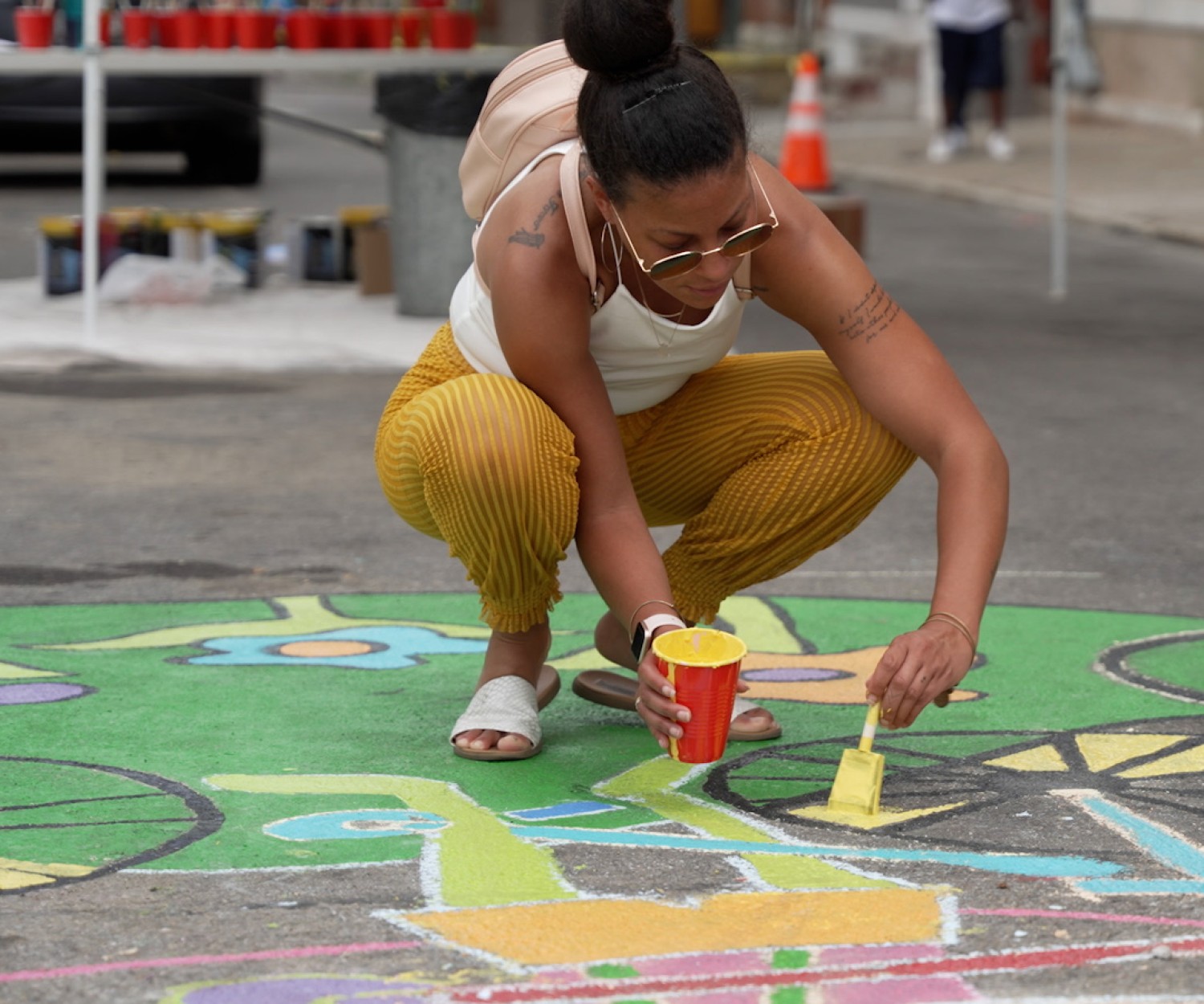 A woman in a white tank top, yellow pants and sandals crouches down, holding a red solo cup in one hand and a paintbrush in the other. She adds a bright yellow paint to a street mural.