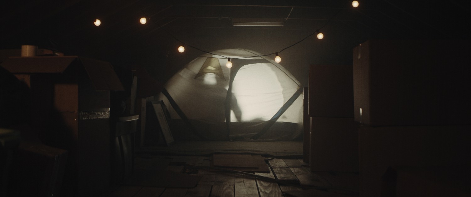A tent sits in the middle of a hazy attic. Boxes surround the tent and string lights dangle in front of it. A light emanates from within the tent and the shadow of a person is seen within.