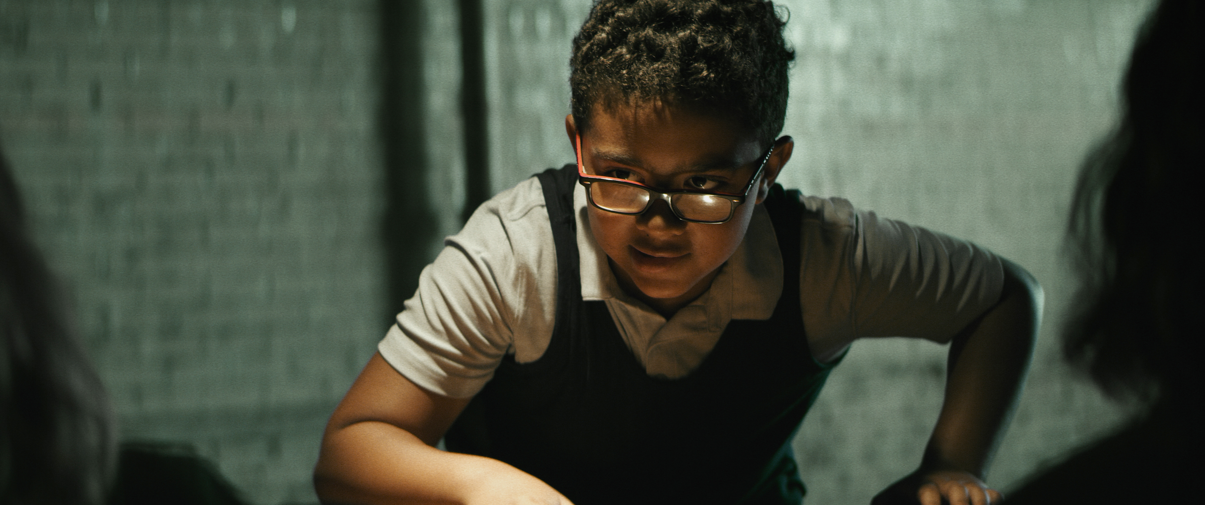 A boy wearing a white polo, black vest and glasses looks intently to the left of frame. He is hunched over a table, hand resting on the table and there are two people out of focus in the foreground.
