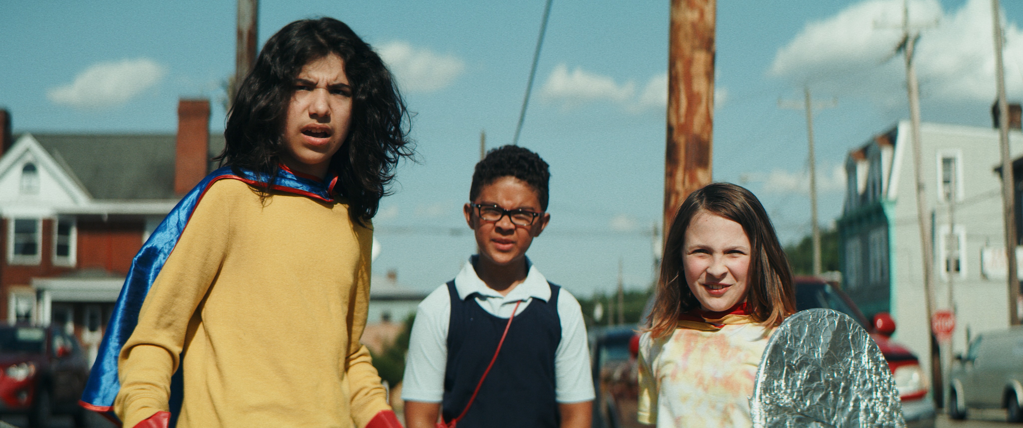 Three kids stand on a city street, side by side, dressed in superhero costumes. All three look forward. The boy on the left wears a yellow shirt and blue cape, while the boy in the center wears a white polo, black vest and glasses. The girl on the right wears a yellow and red tie dye shirt and holds a shield covered in aluminum foil.
