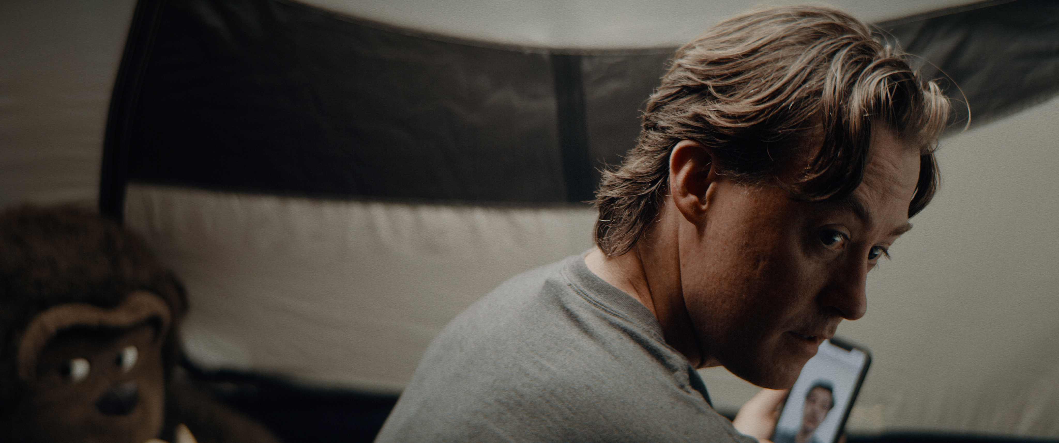 A man sits in a tent, wearing a grey t-shirt. His back is to the camera as he turns his head to look over his right shoulder. He is holding a phone, a man is on the other end of a video call. A stuffed ape sits in the background.