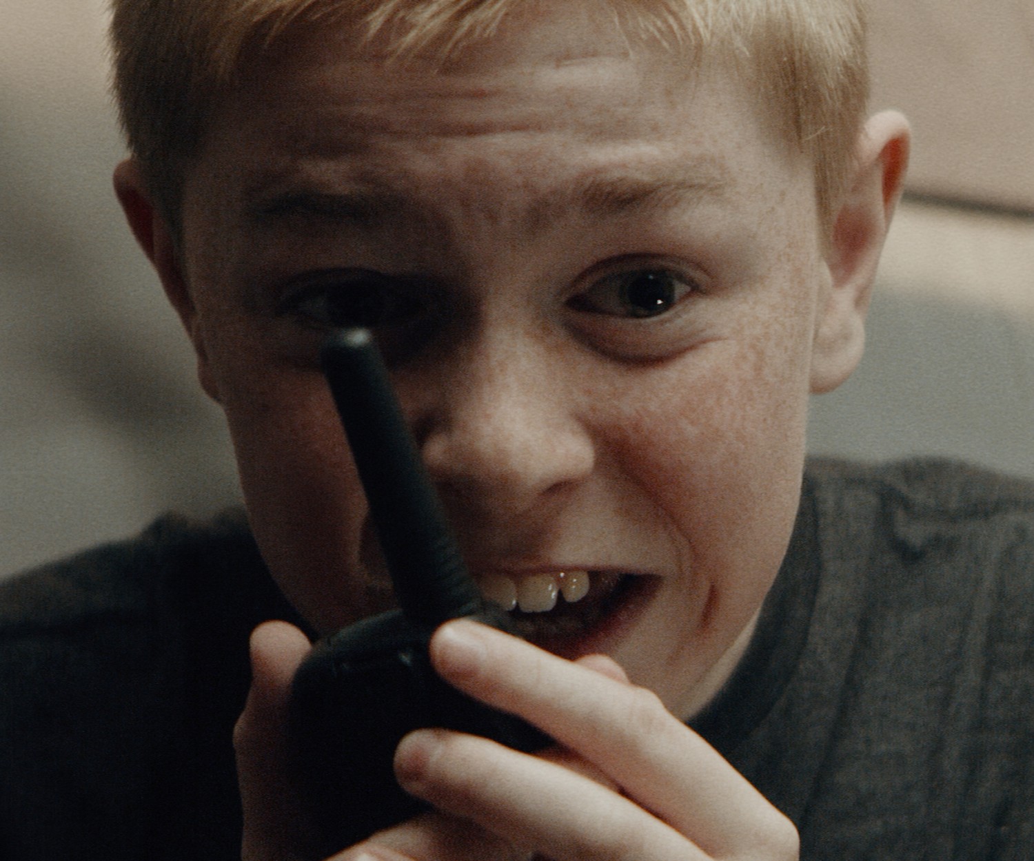 An excited boy sits in a tent, wearing a grey t-shirt. He holds a walkie talkie up to his mouth and is speaking into it.
