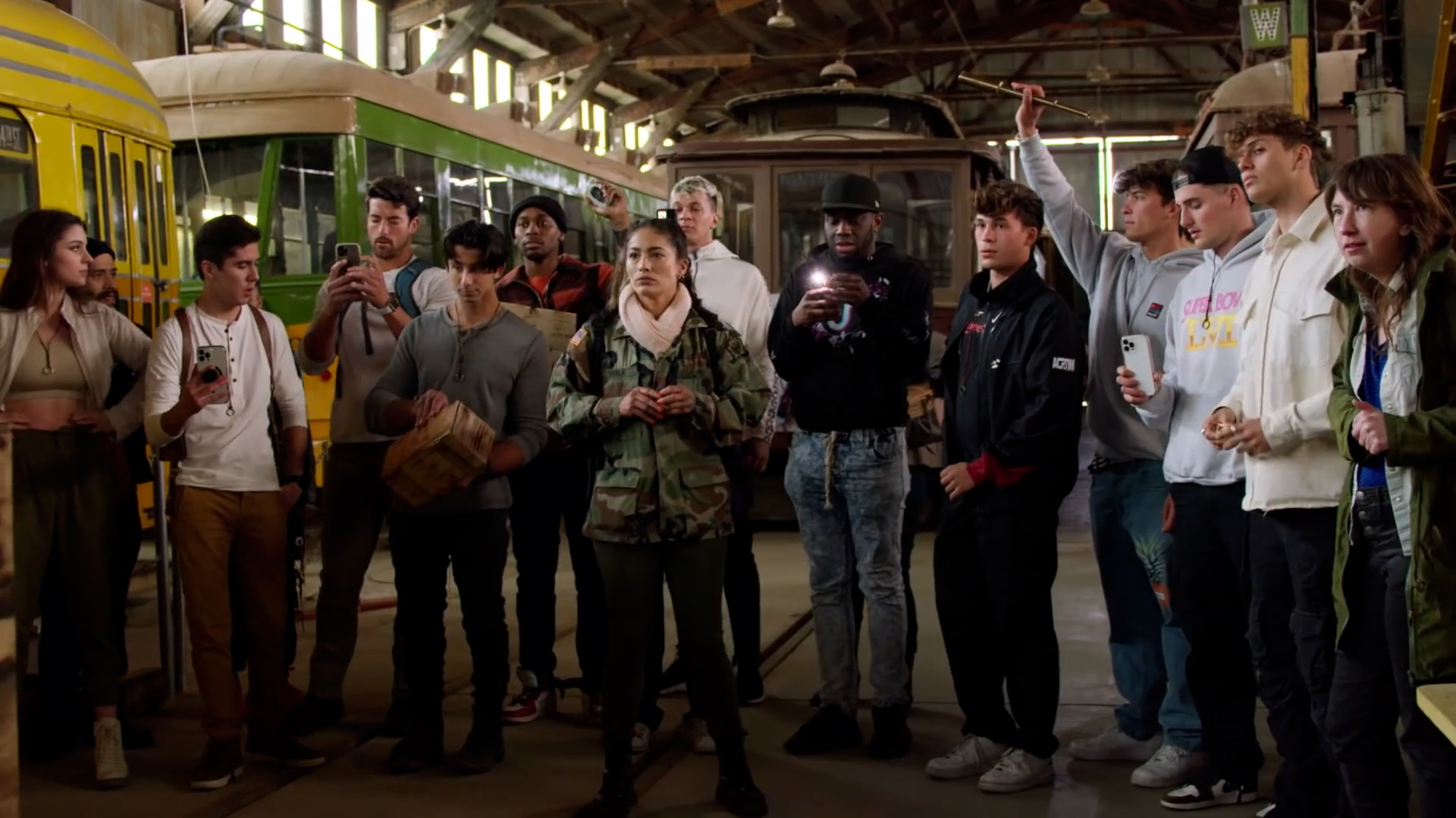 A group of social media influencers stand in a semi-circle, looking forward. Some are holding their phones, recording. They are surrounded by old trolly cars in a dim warehouse.