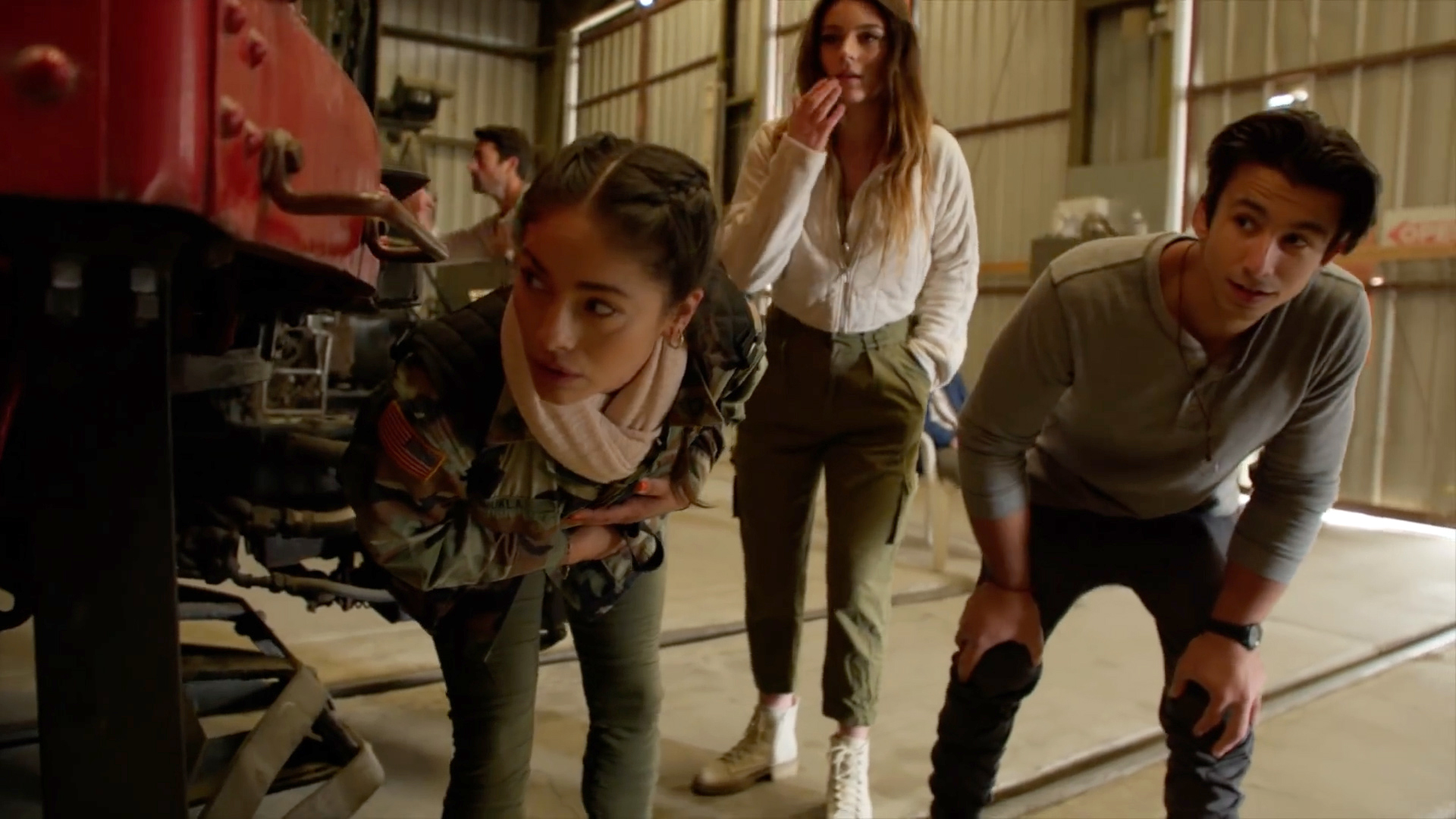 A woman in a camouflage jacket and green pants and a man in a grey raglan and slacks both crouch, peering around the corner of a train car in a dim warehouse. A woman in a white jacket and green pants with white shoes stands behind them.